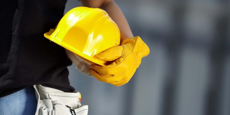 Person in black shirt and blue jeans holding yellow hard hat and wearing protective gloves, with tool belt—emphasizing PPE in construction settings