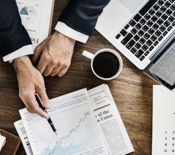A professional workspace showing a person in a suit pointing at a rising financial graph, with a laptop displaying a spreadsheet, coffee cup, planner, and newspaper. The setup reflects strategic planning and financial analysis relevant to renovation project timeline Dubai.