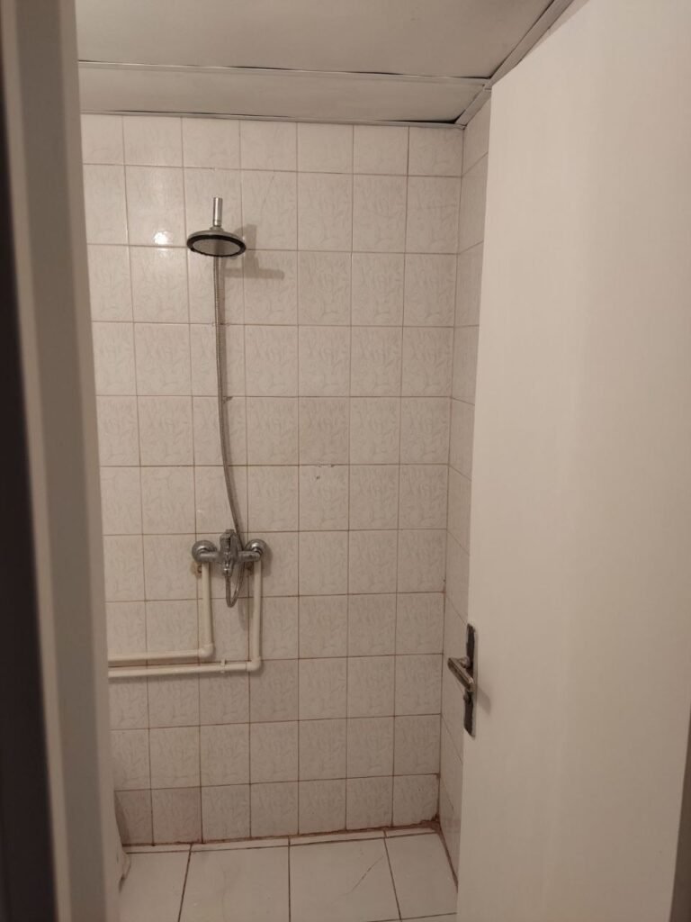 An old shower area with outdated tiles and exposed plastic plumbing pipes before a complete modern renovation by Fardo Technical Services.