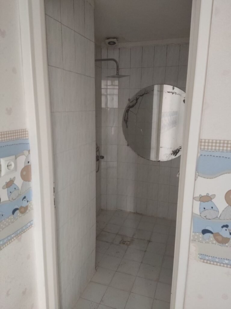 An old outdated bathroom with a damaged round mirror and old white tiles before a complete modern renovation by Fardo Technical Services.